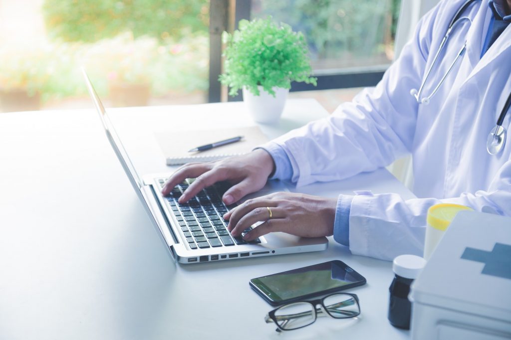 Doctor working with laptop computer and writing on paperwork. Hospital background.