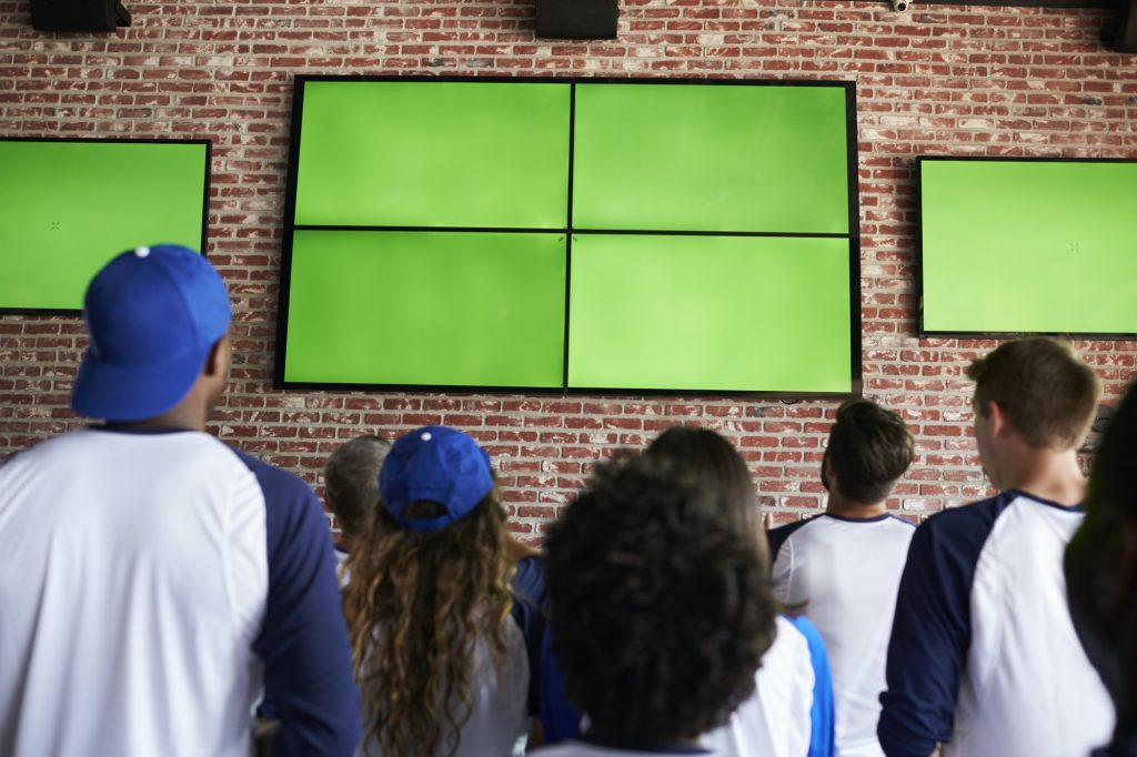 Rear View Of Friends Watching Game In Sports Bar On Screens